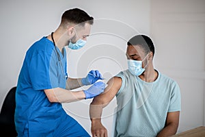 Doctor in face mask giving covid-19 vaccine injection to black male patient during vaccination campaign at clinic