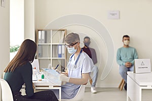 Doctor in face mask interviewing woman and filling out medical record before giving her vaccine