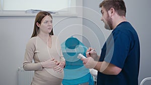 A doctor gives recommendations to a pregnant woman while standing near a gynecological chair. The woman holds her hands