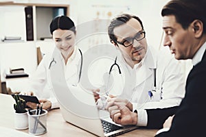 Doctor in glasses advises patient sitting at table with laptop in medical office.