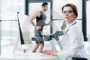 doctor in glasses using computer while sportsman running on treadmill during endurance test in gym.