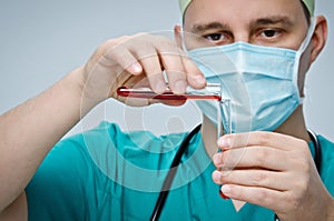 The doctor in a green hat and mask pours red liquid from one test tube to another.