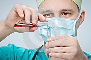 The doctor in a green hat and mask pours red liquid from one test tube to another glass tube.