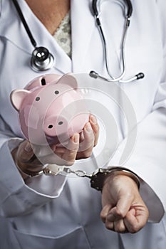 Doctor In Handcuffs Holding Piggy Bank Wearing Lab Coat, Stethoscope