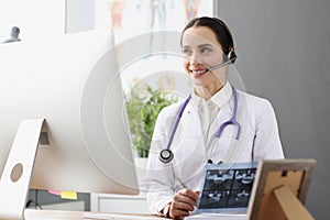 Doctor in headphones with microphone holding x ray in front of computer screen