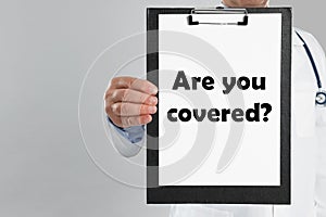 Doctor holding clipboard with text ARE YOU COVERED? on light background. Insurance concept