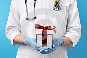 Doctor holding gift box on light blue background, closeup. Medical present