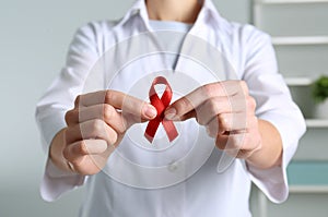 Doctor holding red awareness ribbon indoors, closeup. World AIDS disease day