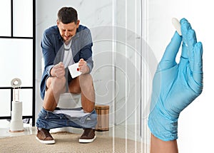 Doctor holding suppository for hemorrhoid treatment and man sitting on toilet bowl in room