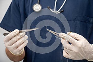 Doctor holds, in hand with disposable glove, forceps and needle holder and suture thread