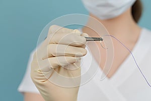 Doctor holds a surgical thread with a needle in gloves with a mask on a blue background