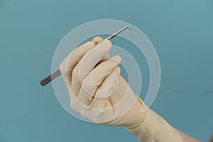 Doctor holds a surgical thread with a needle in gloves with a mask on a blue background