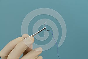 Doctor holds a surgical thread with a needle in gloves with a mask on a blue background