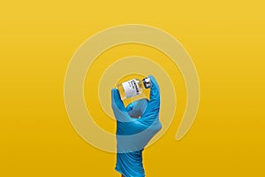 Doctor at laboratory wearing blue rubber glove holding Coronavirus vaccine dose bottle label, isolated on yellow background.