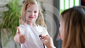 Doctor listens to child with stethoscope, girl holds thumbs up