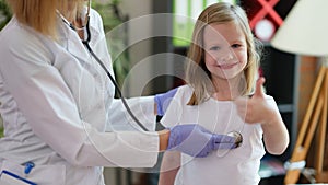 Doctor listens to child with stethoscope girl holds thumbs up