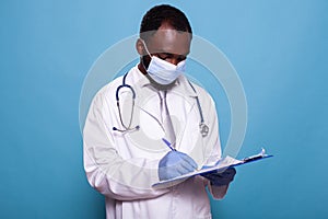 Doctor looking down at clipboard with patient charts wearing lab coat, surgical mask and latex gloves