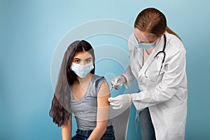 Doctor making injection of coronavirus vaccine to Indian teenage girl in face mask on blue studio background