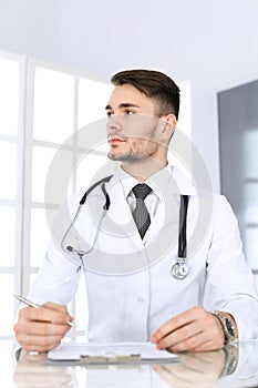 Doctor man filling up medication history records form while sitting at the glass desk in medical office or clinic