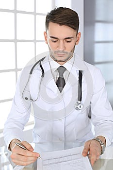 Doctor man filling up medication history records form while sitting at the glass desk in medical office or clinic