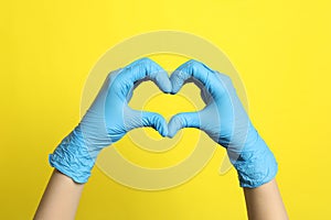 Doctor in medical gloves making heart with hands on yellow background, closeup