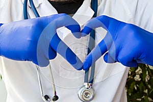 Doctor in medical lab coat with stethoscope shows shape of card heart close-up in front, made using fingers, wearing in blue glove