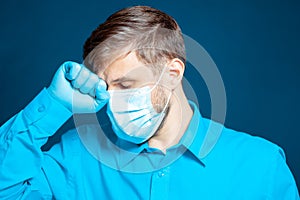 doctor in a medical mask and gloves, in a blue uniform, leaned his fist against his head