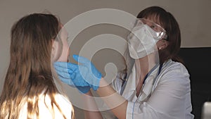 A doctor in medical mask with goggles and medical gloves examines throat and eyes of child. woman in white coat and