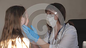 A doctor in medical mask with goggles and medical gloves examines throat and eyes of child. woman in white coat and