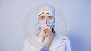 A doctor in a medical mask holds a finger in front of his mouth, a gesture of silence or secretion.