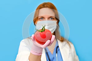 Doctor nutritionist in a medical mask with a red tomato in his hand on a blue background, portrait