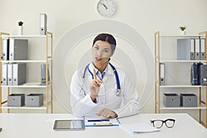 Doctor online. Hispanic female doctor looking at the camera says diagnose while sitting at a table in a clinic office