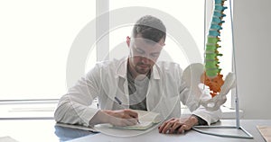 Doctor orthopedic surgeon is doing his paperwork sitting in his office with spine model on the table.