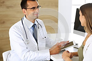 Doctor and patient discussing medical exam results while sitting at the desk in clinic. Male physician using tablet