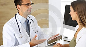 Doctor and patient discussing medical exam results while sitting at the desk in clinic. Male physician using tablet