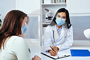 Doctor and patient in face masks discussing health problems while sitting at the table in medical center