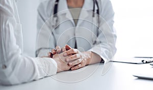 Doctor and patient sitting at the desk in clinic office. The focus is on female physician& x27;s hands reassuring woman