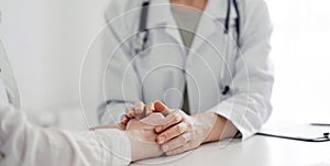 Doctor and patient sitting at the desk in clinic office. The focus is on female physician& x27;s hands reassuring woman