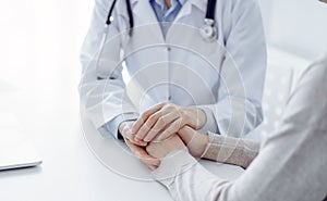 Doctor and patient sitting at the table in clinic office. The focus is on female physician& x27;s hands reassuring woman