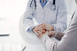 Doctor and patient sitting at the table in clinic office. The focus is on female physician& x27;s hands reassuring woman