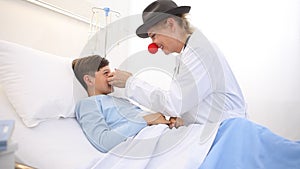 Doctor pediatrician clown with red nose and happy child patient lying in hospital bed, cheers up loneliness with alternative