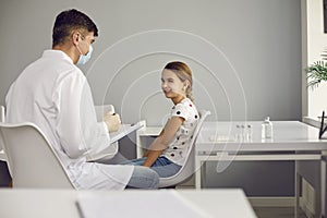 Doctor pediatrician in medical uniform and mask talking with little smiling girl patient