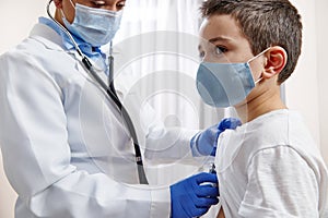 Doctor pediatrician in medical uniform uses a stethoscope while auscultating a little boy in protective medical mask photo