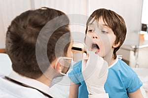 Doctor in protective mask and gloves examining throat of child