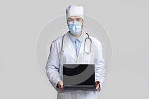 Doctor in protective mask showing laptop with blank screen