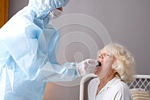 Doctor in protective suit taking throat and nasal swab from elderly female patient to test for infection