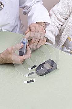 Doctor punches the finger of a very old woman. The glucometer with stripes lies on the table.