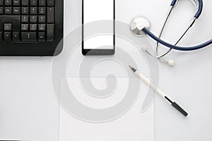 A doctor`s desk viewed from above with a computer keyboard, a smartphone with blank screen, a stethoscope and a notebook for