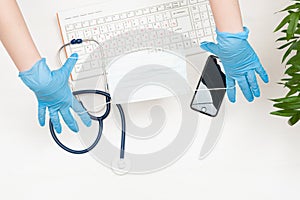 The doctor`s gloved hands hold the mask. Keyboard, stethoscope, phone, mask, gloves. Top view.