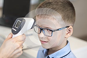 Doctor`s hand measures the temperature of the boy in a blue shirt and glasses using an electronic infrared thermometer.